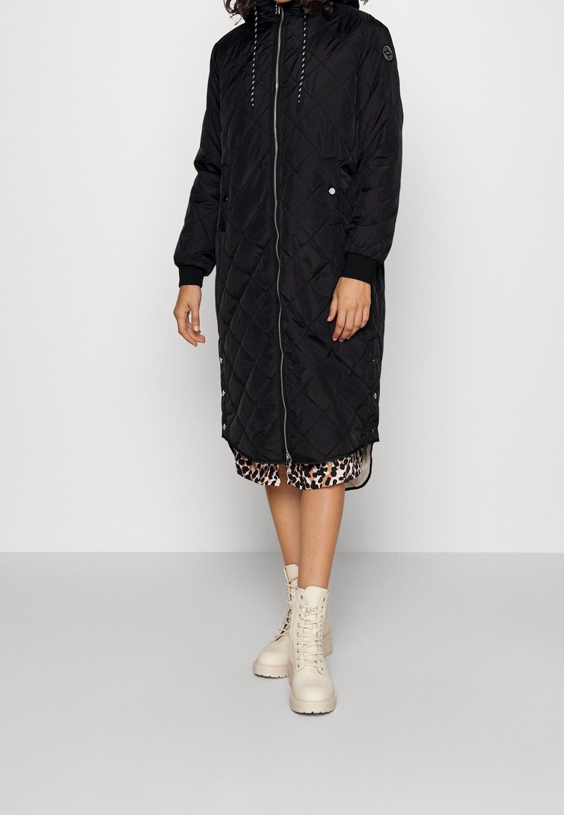 Abrigo largo acolchado negro con capucha, cierre de cremallera y detalles a presión en los laterales. Llevado sobre un vestido de estampado de leopardo y combinado con botas blancas de cordones.