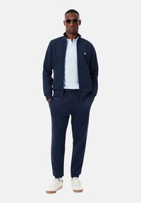 Temno modra trenirka z jopico z zadrgo in hlačami z elastičnim pasom, v kombinaciji s svetlo modro polo majico in belimi supergami z bež podplati.