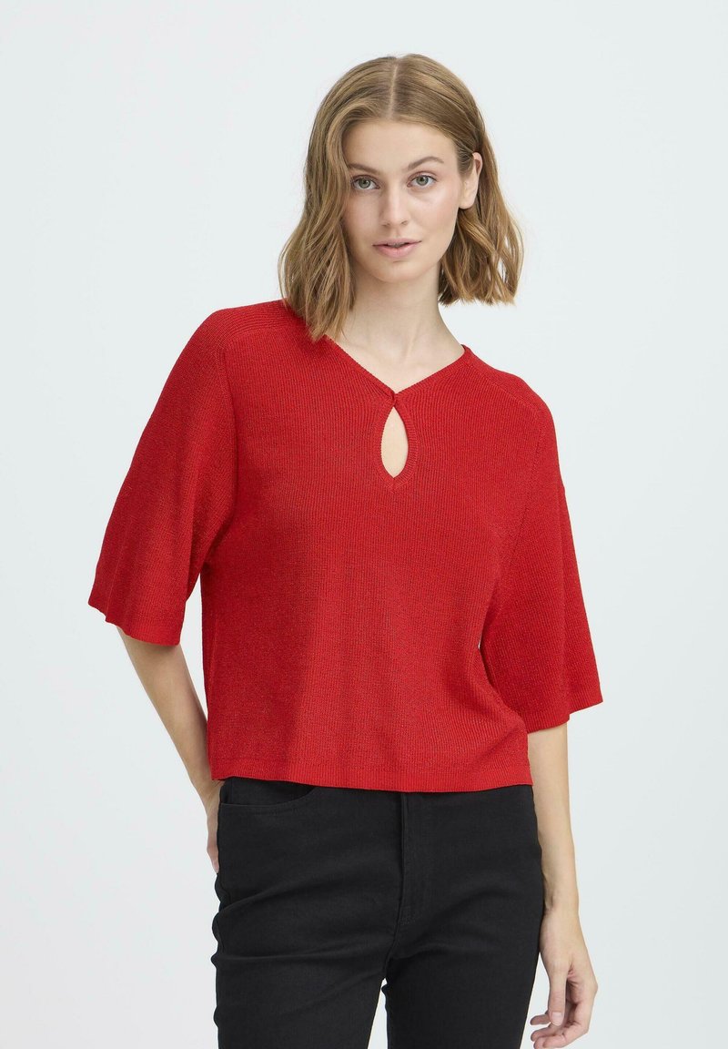 Crvena pletena majica s V-izrezom i detaljem u obliku ključa, s kratkim rukavima i opuštenim krojem, u kombinaciji s crnim hlačama.