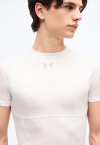 Νεαρός άνδρας που φοράει εφαρμοστή λευκή συμπιεστική κοντομάνικη μπλούζα με το λογότυπο Under Armour στο στήθος, κοιτάζοντας προς τα δεξιά.