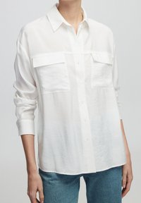 Chemise blanche à manches longues avec deux poches poitrine, un col classique et une coupe légèrement ample. Tissu texturé.