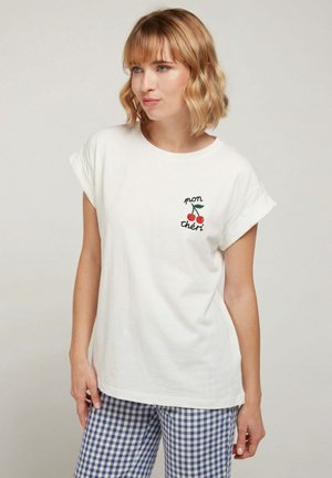 Jeune femme aux cheveux blonds mi-longs portant un T-shirt blanc avec un motif de cerise et le texte « mon chéri », et un pantalon à carreaux bleu.