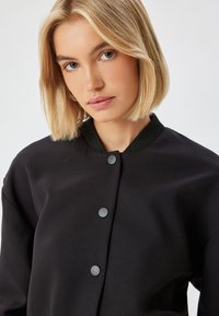 Mustat pommitakkia, jossa on tekstuuripinta, ribbireunainen kaulus ja neljä harmaata nepparia edessä. Yksinkertainen ja moderni muotoilu.