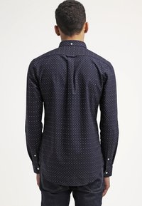 Granatowa koszula zapinana na guziki w białe kropki, z długim rękawem i zakładką na plecach. Posiada klasyczny kołnierzyk i mankiety zapinane na guziki.