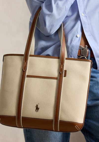 Personne portant une chemise bleue et un jean tenant un sac cabas beige et marron avec garniture en cuir et logo Polo Ralph Lauren.