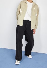 Persona vestida con chaqueta beige con cremallera, camisa blanca, jeans negros de pierna ancha y zapatillas blancas con patrones negros, de pie sobre un suelo azul.