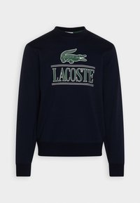 Tmavě modrá mikina vyrobená z bavlny, s kulatým výstřihem, dlouhými rukávy a zeleným krokodýlem s nápisem "LACOSTE" pod ním v bílém písmu.