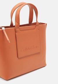 Borsa a mano in pelle arancione con manici doppi. Presenta una tasca piatta con logo impresso. Texture liscia e design strutturato. Chiusura con zip.