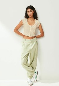 Top court beige avec motif en losange et manches à volants, associé à un pantalon cargo vert clair et des baskets blanches avec des accents verts.