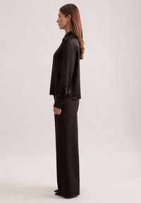 Donna in blusa nera di raso a maniche lunghe e pantaloni larghi, in profilo su uno sfondo chiaro e semplice.