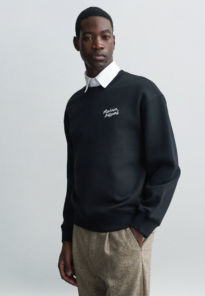 Čierna mikina s rebrovanými manžetami a spodným lemem, s bielym vyšívaným textom. Pod ňou je vidieť bielu košeľu s golierom.