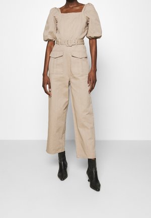 Beżowy lniany kombinezon z bufiastymi krótkimi rękawami, kwadratowym dekoltem, paskiem w talii i dwoma przednimi kieszeniami. Połączony z czarnymi, szpiczastymi botkami za kostkę.
