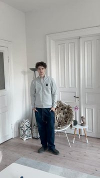 Pilkas zip-up megztinis, mėlynos kelnės ir tamsi avalynė. Kambaryje šviesios sienos, medinis grindys, augalai ir dekoratyviniai daiktai.