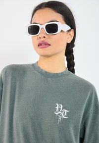Grå oversized t-shirt med et hvidt grafisk logo på brystet. Modellen bærer hvide firkantede solbriller og har en fletning.