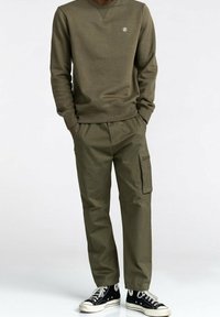 Felpa verde oliva con scollo tondo e piccolo logo, abbinata a pantaloni cargo verde oliva con tasche laterali e sneaker nere.