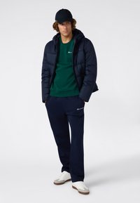 Giacca puffer navy con cappuccio sopra un sweatshirt verde, abbinata a pantaloni navy e sneakers bianche. Presenta un logo sulla felpa e sui pantaloni.