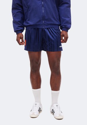 Personne portant un short de sport Adidas bleu marine, une veste assortie, des chaussettes blanches montantes et des baskets Adidas blanches avec des rayures noires.