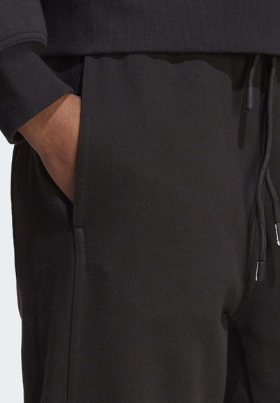 Schwarze Jogginghosen mit elastischem Bund aus glattem Material, ausgestattet mit Seitentaschen und einer Kordelzugvorrichtung. Nahaufnahme des Taschendetails.