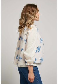 Veste en polaire blanche texturée avec des motifs floraux bleus, manches cloche et ourlet arrondi. Portez-la sur une blouse légère et associez-la à un jean bleu.