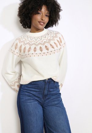 Kreminė megzta palaidinė su geometriniu raštu rožinės spalvos ir metaliniais akcentais kaklo srityje, dėvima su aukštu liemeniu tamsiai mėlynomis džinsais.