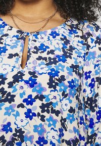 Bloes met bloemenprint in blauw en wit, met een keyhole halslijn en zachte stof. Inclusief gelaagde gouden kettingen.