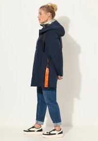 Marinblå huva jacka med en glänsande silvrig innerfoder, två framfickor, orange dragkedjedetalj och svarta remaccent. Påsatt över denimjeans.