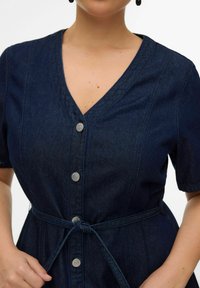 Mujer con vestido de mezclilla azul oscuro de manga corta, botones plateados al frente y cinturón atado a la cintura.