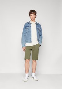 Giubbotto di jeans azzurro chiaro, polo bianca, pantaloni corti verde oliva e sneakers bianche con dettagli grigi. Modello in piedi contro uno sfondo neutro.