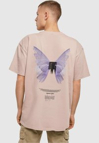 Kortærmet, pastelpink t-shirt med et stort lilla sommerfugletryk på ryggen, med tekst og designelementer nedenfor.