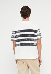 Λευκό μπλουζάκι με μαύρες οριζόντιες ρίγες στην πλάτη. Κοντά μανίκια με άνετη εφαρμογή και στρογγυλή λαιμόκοψη. Απαλή υφή από βαμβάκι.