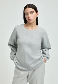 Grauer Pullover aus weichem Stoff, mit Rundhalsausschnitt, überschnittenen Schultern und langen Ärmeln. Kombiniert mit karierten Hosen.