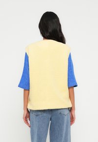 Veste sans manches en polaire jaune à finition texturée, associée à une chemise à manches courtes bleue, portée sur un jean délavé clair.