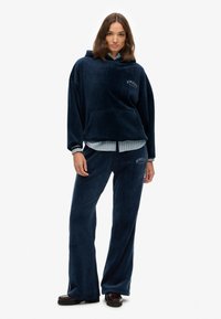 Sudadera de terciopelo azul marino con un bolsillo frontal y texto bordado. Combinada con pantalones de pierna ancha a juego, con una camisa a rayas por debajo.