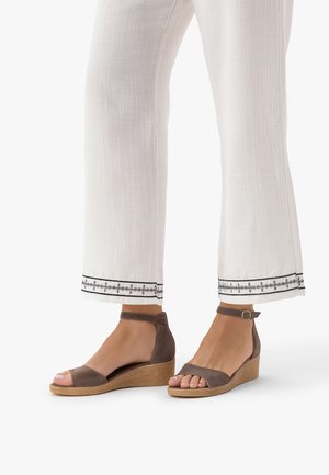 Nogi w białych spodniach z czarnym haftowanym wykończeniem oraz brązowych zamszowych sandałach na koturnie z paskami wokół kostki, widok z boku.