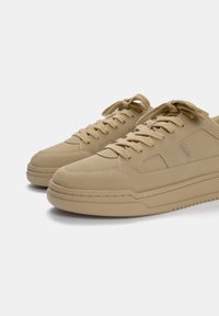 Zapatilla beige que presenta una parte superior sintética lisa, puntera redonda, cierre de cordones y suela texturizada con un detalle sutil de logotipo en el lateral.