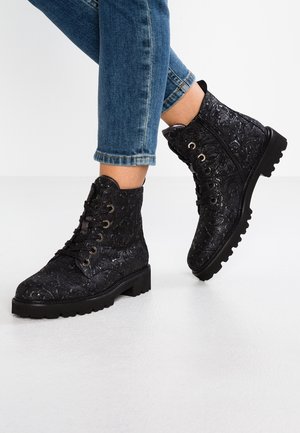 Bottes noires à lacets embossées avec fermeture éclair latérale, portées avec un jean bleu retroussé sur un sol gris clair.
