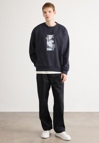Felpa navy con un design grafico sul davanti, abbinata a pantaloni neri e sneakers bianche. Materiale in cotone morbido e vestibilità rilassata.