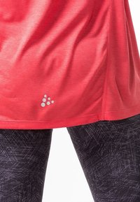 Röd idrottströja med en mjuk textur, med en liten reflekterande logotyp nära fållen. Kombinerad med svarta mönstrade leggings.