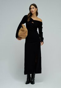 Schwarzes langärmliges Kleid mit asymmetrischem Ausschnitt und seitlicher Schlitze. Das Model hält eine hellbraune übergroße Tasche und trägt schwarze kniehohe Stiefel.