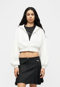 Γυναίκα με μακριά σκούρα μαλλιά, που φοράει λευκό κοντό φούτερ με φερμουάρ, μαύρο αθλητικό τοπ και μαύρη πλισέ φούστα με λογότυπο Puma.