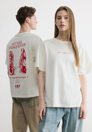 Egy nő szemből néz, fehér pólót visel, egy férfi oldalról áll, és fehér pólója hátulját mutatja piros sportcipő grafikával és "Nem a szokásos futó" felirattal.