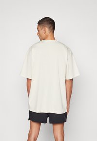 Hombre con el cabello corto que lleva una camiseta blanca oversized y pantalones cortos negros, de pie de espaldas contra un fondo liso.
