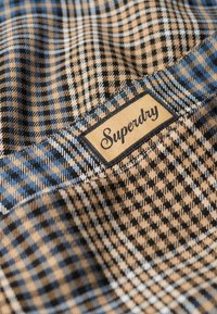 Tissu à carreaux marron et bleu avec un motif texturé, comportant une étiquette tissée avec l'inscription « Superdry » en écriture cursive sur un fond beige.