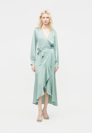 Mujer con cabello rubio que lleva un vestido cruzado de manga larga color verde claro, con cintura atada y bajo asimétrico con volantes, posando frente a un fondo liso.