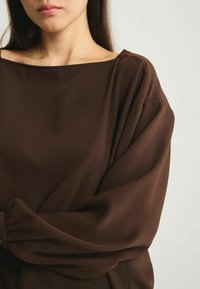 Blouse marron fluide avec un large col bateau et des manches longues froncées, fabriquée dans un tissu léger, présentant une texture lisse.