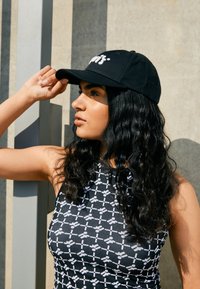 Melnā cepure ar izšūtu logo, ko valkā cilvēks ar gariem viļņainiem matiem. Cepures augšdaļai ir izliekts maliņš, un fona attēlā ir teksturēta siena.