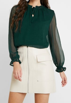 Vrouw draagt een donkergroene transparante blouse met lange mouwen en ruches aan de manchetten en kraag, gecombineerd met een witte hoge taille minirok met een rits aan de voorkant en een zak.