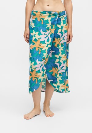 Personne pieds nus portant une jupe portefeuille fleurie colorée avec des motifs bleus, verts, oranges et jaunes, debout devant un fond uni.
