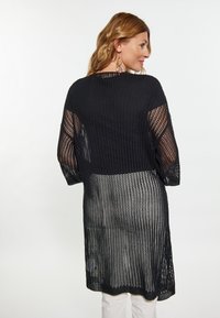 Fekete, nyitott kötésű kardigán bő, rövid ujjal; áttetsző textúra; oversize fazon; a hem a comb közepéig ér; fehér nadrággal párosítva.
