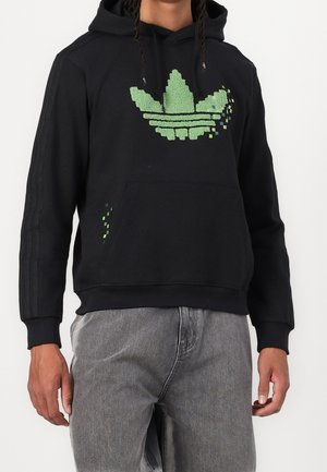 Mann trägt schwarzen Hoodie mit pixeligem grünem Blätter-Logo und graue Hose, steht vor weißem Hintergrund.